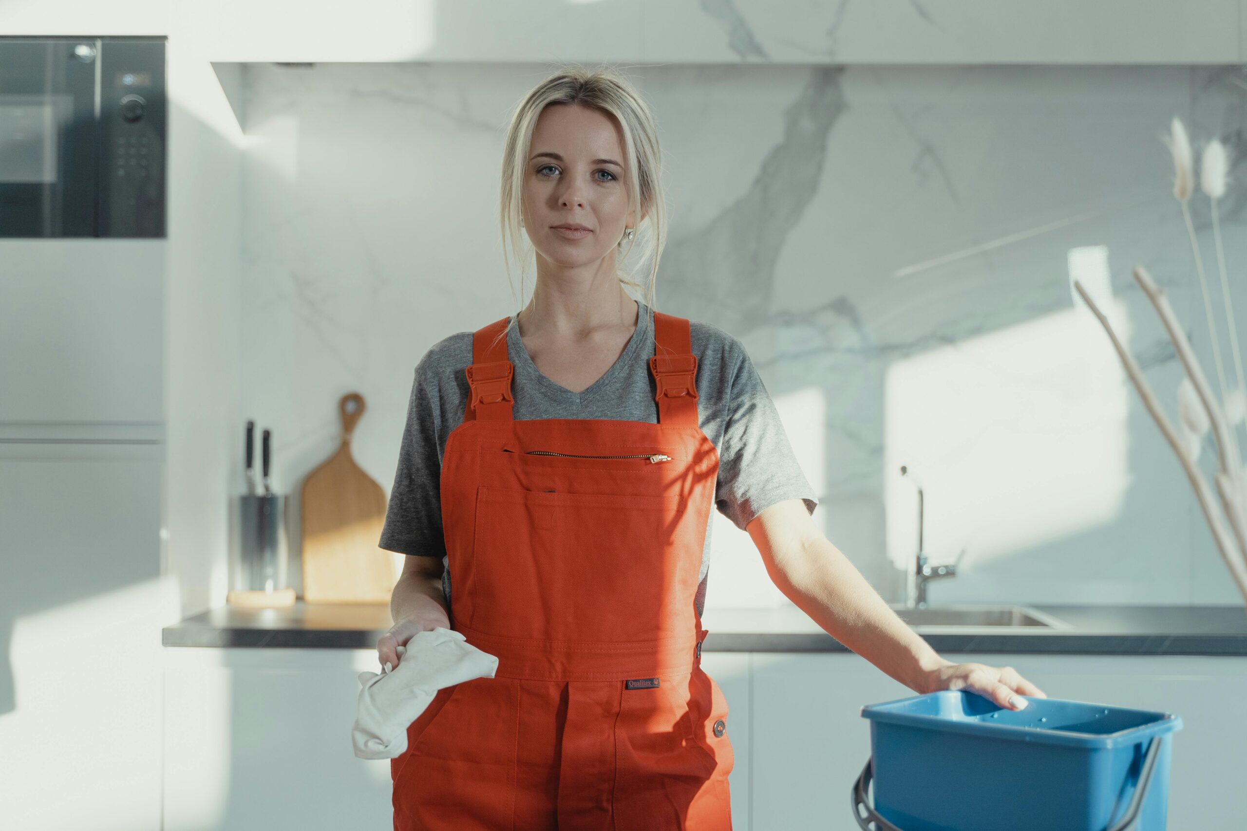 A woman in an orange apron stands in a modern kitchen holding a cleaning cloth next to a bucket.