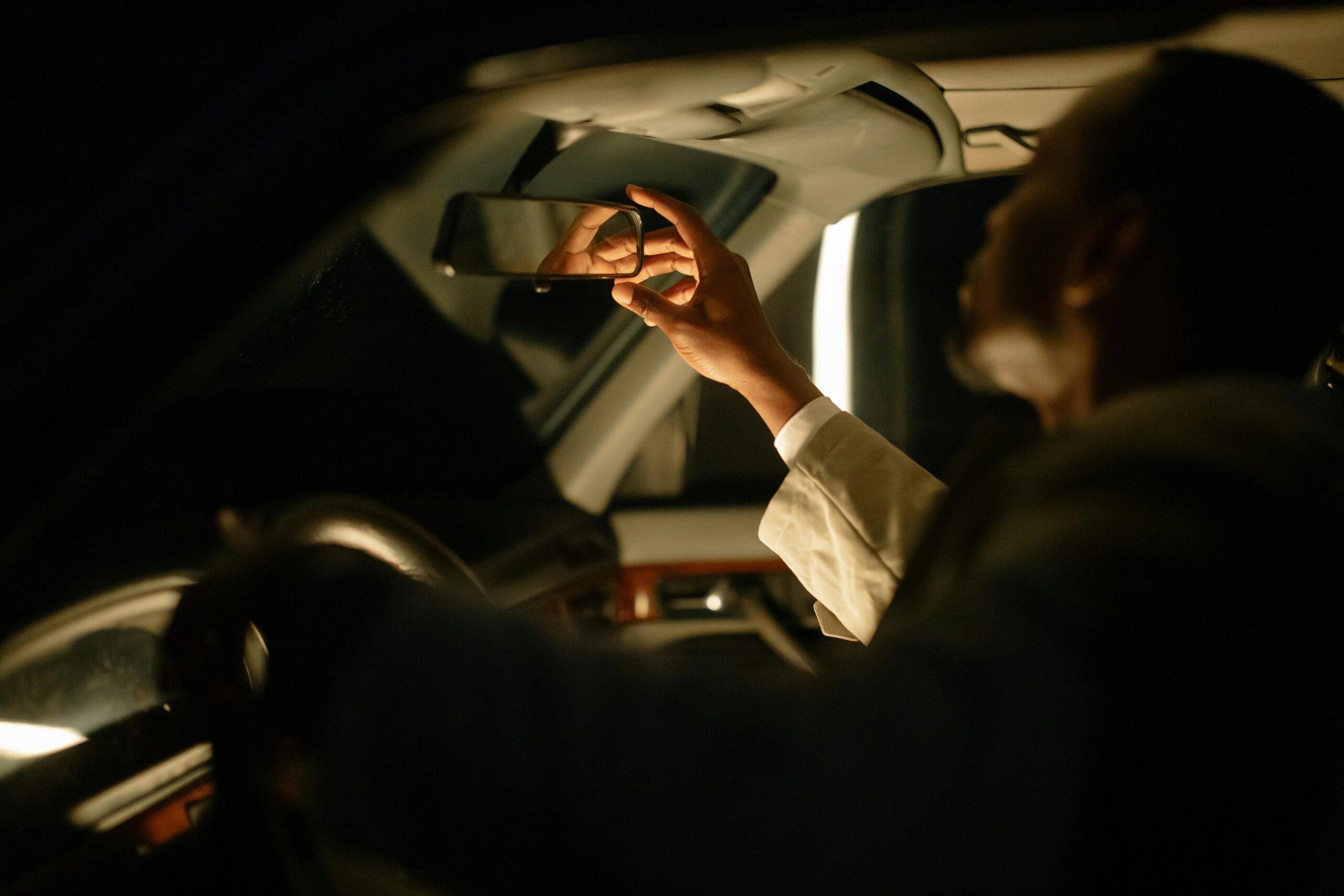 A man adjusting the rear view mirror inside a car, captured during nighttime with soft lighting.